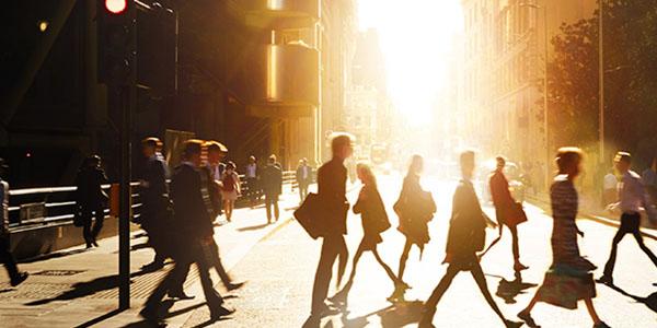 Unscharfes Bild von Passantinnen und Passanten, die eine Straße überqueren mit Gebäuden im Hintergrund und blendendem Sonnenlicht von vorne zwischen den Gebäuden